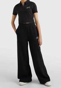 Černé zkrácené polo triko se zipem a vyšívaným logem, kombinované s širokými černými tepláky. Obě z měkké látky. Viditelné bílé tenisky.