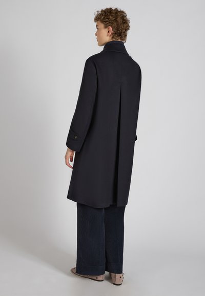 Granatowy płaszcz o długim kroju, z wykładanym kołnierzem i plisą z tyłu. Posiada zapinane na guziki mankiety i jest zestawiony z szerokimi spodniami dżinsowymi.
