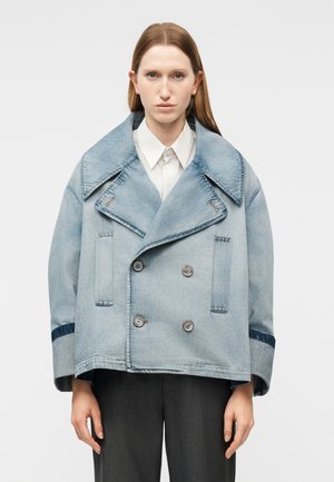 Femme portant une veste oversize en denim bleu clair à double boutonnage par-dessus une chemise blanche et un pantalon foncé, debout devant un fond uni.