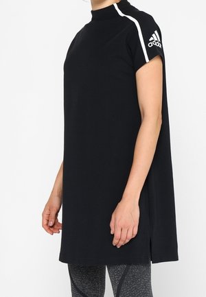 Vrouw die een zwarte Adidas-tuniek draagt met witte strepen op de mouwen, gecombineerd met grijze legging met patroon, zijwaarts staand.