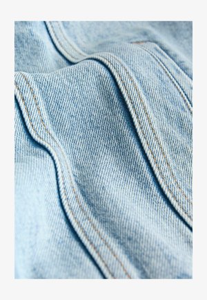 Svetlomodrá rifľovina s viditeľnou textúrou, s vlnitými záhybmi a kontrastným oranžovým stehom pozdĺž švov.