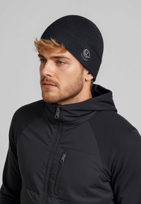 Czarna dzianinowa czapka z gładkim wzorem, z małym logo z boku. Noszona z czarną kurtką na zamek błyskawiczny.