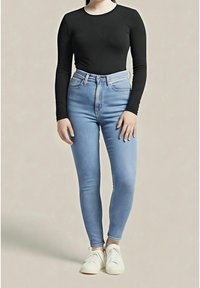 Mujer con un top negro de manga larga, jeans ajustados de tiro alto en azul claro y zapatillas blancas, de pie contra un fondo liso.