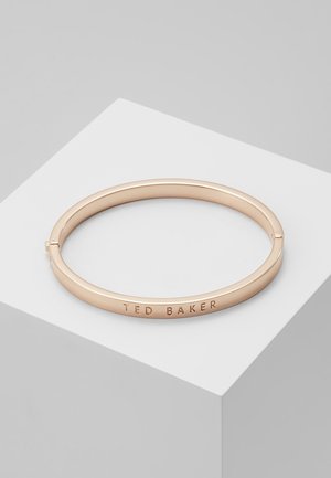 Rosa guld cirkulær armring med en glat finish, graveret med "TED BAKER," hvilende på en hvid skrå flade.