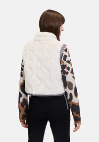 Kurze, cremefarbene Kunstfellweste mit gestepptem Muster, kombiniert mit einem gemusterten Langarmshirt in Braun-, Schwarz- und Beigetönen; schwarze Hose.