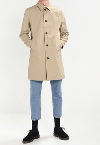 Trench coat di un beige chiaro con colletto, chiusura frontale a bottoni e tasche laterali, abbinato a jeans cropped azzurri e scarpe nere.