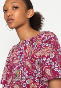 Blusa de padrão paisley roxo com mangas curtas, apresentando enfatizações florais em branco e laranja. Tecido suave com decote redondo.