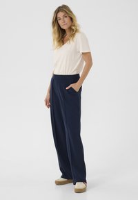 T-shirt crème à col en V, associé à un pantalon large marine avec poches latérales. Le mannequin se tient sur une surface lisse, portant des chaussures beiges.