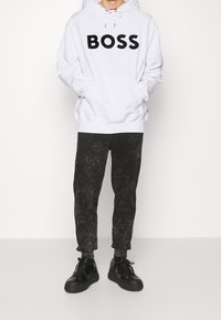 Weißer Hoodie mit schwarzem "BOSS"-Logo, kombiniert mit dunkel marmorierten Jogginghosen und schwarzen Sneakers mit goldenen Akzenten. Einfaches Design und lässige Passform.