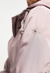 Jasnoróżowa kurtka z warkoczowym sznurkiem i haftowaną gwiazdą na rękawie, wykonana z gładkiego, wodoodpornego materiału.