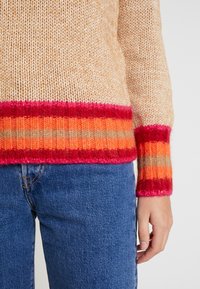 Stickad tröja i beige med en strukturerad yta, med breda randiga manschetter i röd, orange och rosa. Kombinerad med blå denimbyxor.