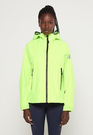 Frau trägt eine leuchtend neon-grüne Kapuzenjacke mit schwarzem Reißverschluss und marineblaue Leggings, steht vor einem schlichten hellen Hintergrund.