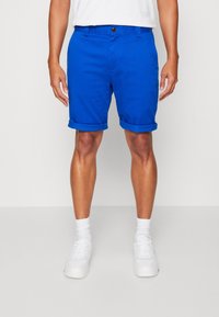 Blå bomullsshorts med uppvikta ärmar, med knappstängning och framfickor, ihop med vita strumpor och sneakers.