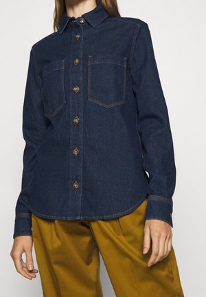 Kvinde iført en mørkeblå denimskjorte med brystlommer og sennepsgule bukser, stående mod en ensartet hvid baggrund.