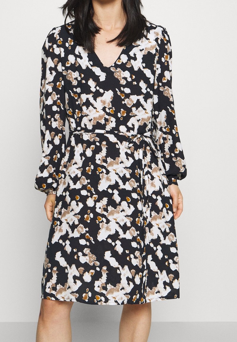Femme portant une robe à manches longues au motif abstrait noir, blanc et marron, avec un col en V et une ceinture nouée à la taille.