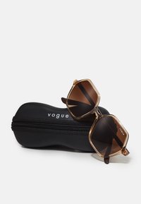 Hexagonal brown sunglasses with a transparent tan frame, resting on a black zippered case featuring the text "vogue" in white.