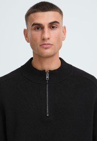 Schwarzer Strickpullover mit hohem geripptem Kragen und halbem Reißverschluss, der eine strukturierte Oberfläche und eine figurbetonte Silhouette aufweist.