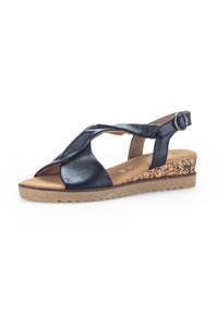 Sandalia de cuero negro con diseño de tiras cruzadas, punta abierta y hebilla en el tobillo. Presenta un tacón tipo cuña texturizado similar al corcho y suela de goma.