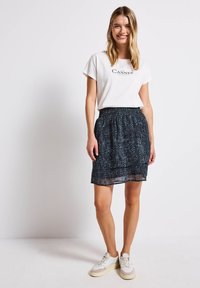 Weißes T-Shirt mit "Cannes"-Aufdruck, kombiniert mit einem schwarz-blauen gemusterten Rock und weißen Sneakers. Der Rock hat einen elastischen Bund.
