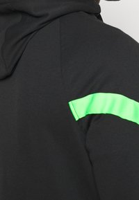 Haut à capuche noir avec une texture lisse et une bande horizontale verte éclatante sur le haut du bras. Le tissu semble léger et sportif.