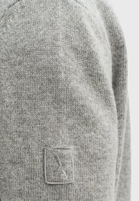 Szary sweter z dzianiny o gładkiej fakturze, z małym haftowanym logo na prostokątnej łatce na ramieniu.