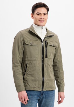 Olive green jacket with zip front, two chest pockets, and side pockets; fabric texture appears smooth. Worn over a white high-neck top.