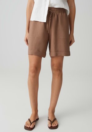 Vrouw draagt bruine knielange shorts met opgerolde zoom, voorzakken, aantrekkoord in de taille, wit topje en bruine slippers, staand op een grijze vloer.