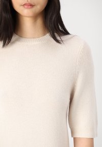 Beiger Strickpullover mit rundem Halsausschnitt und kurzen Ärmeln, der eine feine Textur und gerippte Ärmelabschlüsse aufweist.