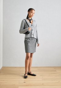 Cardigan en tricot gris avec col en V et boutons, associé à une jupe ajustée à carreaux. Des ballerines noires et une écharpe à motifs noir et blanc viennent compléter la tenue.