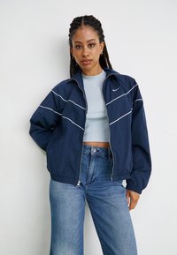 Jovem mulher com cabelo trançado, vestindo uma jaqueta Nike azul marinho, um top curto azul claro e jeans azuis de cintura alta, encostada a uma parede branca.