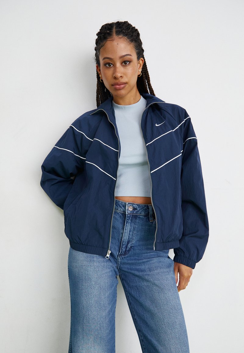 Jeune femme avec des cheveux tressés portant une veste Nike bleu marine, un haut court bleu clair et un jean bleu taille haute se tenant contre un mur blanc.