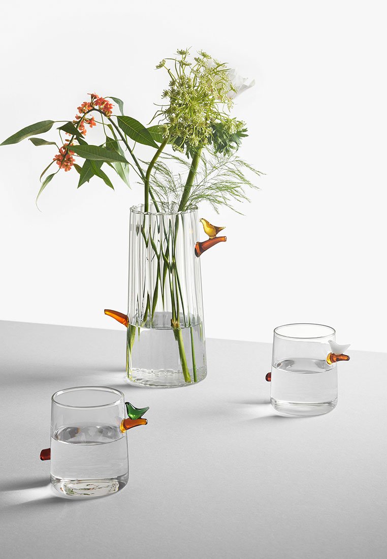 Przezroczysty szklany wazon z pionowymi żłobieniami, wypełniony zielonymi roślinami i kwiatami. Dwa przezroczyste kieliszki z kolorowymi akcentami w kształcie ptaków na uchwytach.