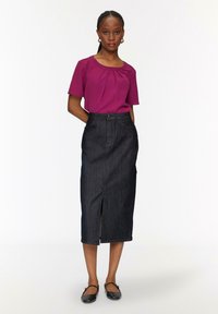 Paarse top met korte mouwen met plooidetail, gecombineerd met een donkerdenim kokerrok met een split aan de voorkant en knoopsluiting. Zwarte platte schoenen.