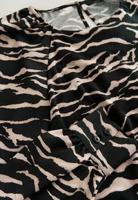 Blusa negra y beige con un patrón de rayas de tigre, que presenta una textura suave y un detalle de puño abotonado. Escote redondo con un corte en forma de llave.