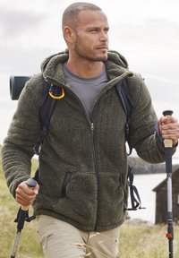 Olivengrüner Fleece-Jacke mit Reißverschluss, zwei Seitentaschen und Kapuze. Ausgestattet mit einem schwarzen Rucksack und Trekkingstöcken.