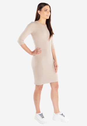 Kobieta z długimi brązowymi włosami, ubrana w beżową sukienkę do kolan i białe trampki, stoi z jedną ręką na biodrze.