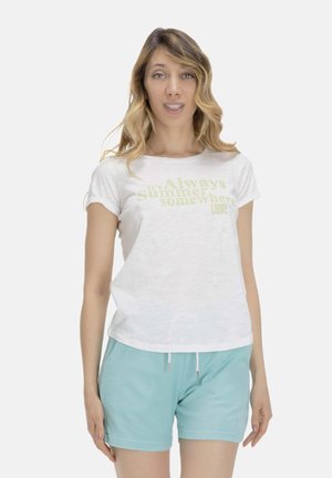 Femme aux cheveux blonds ondulés portant un t-shirt blanc avec un texte clair et un short bleu clair à cordon, sur un fond uni.