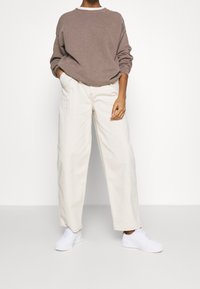 Brun oversized sweatshirt, krämfärgade vida byxor och vita sneakers. Synlig sömnad och avslappnad passform genomgående. Minimalistisk design.