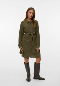 Robe chemise vert olive avec un devant boutonné, des manches longues et une ceinture à la taille, accompagnée de bottes en cuir marron hauteur genoux.