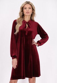 Burgundy bársony ruha hosszú ujjakkal, nyakán kötővel és harang alakú fazonnal. Simább textúra és térd fölé érő fazon.