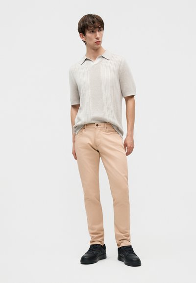 Νέος άνδρας φοράει ένα ανοιχτό μπεζ πλεκτό πόλο με κοντά μανίκια, μπεζ παντελόνι και μαύρα αθλητικά παπούτσια, στέκεται μπροστά σε ένα απλό λευκό φόντο.