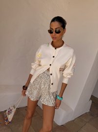 Krémový pletený cardigan s gombíkovým zapínan�ím, doplnený o lesklé strieborné šortky zo揺pôlivých flitrov. Doplnky zahŕňajú slnečné okuliare a korálkový náramok.