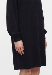 Femme portant une robe noire à manches longues, longueur genou, avec des poignets côtelés, debout devant un fond uni clair.
