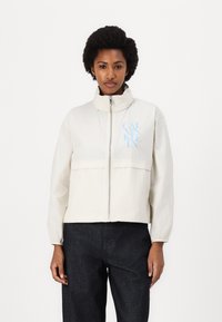 Light cream zip-up jacket with a high collar, featuring a front pocket and blue logo on the left side. Worn with dark denim pants.
