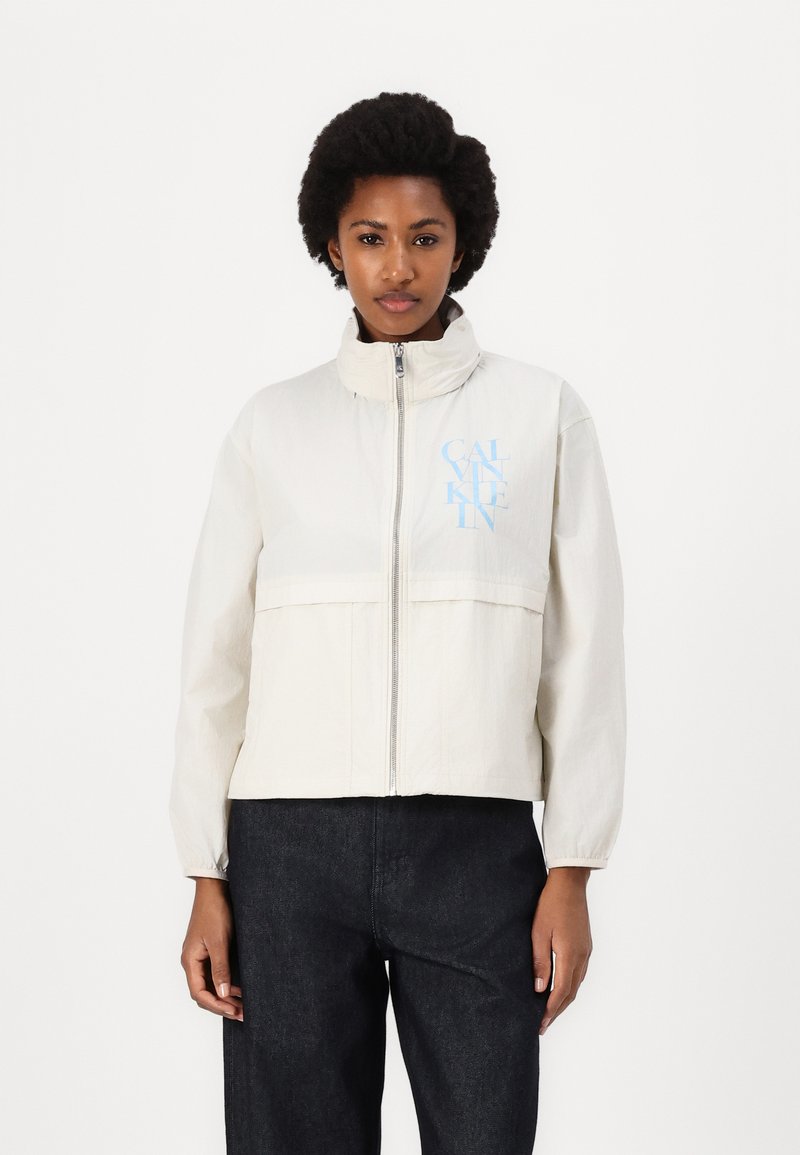 Light cream zip-up jacket with a high collar, featuring a front pocket and blue logo on the left side. Worn with dark denim pants.