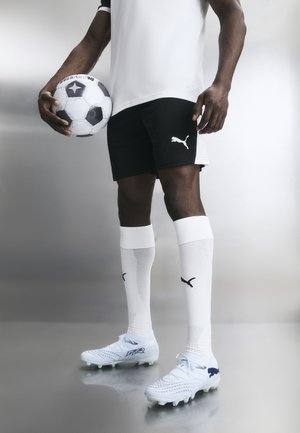Férfi fekete-fehér focimezben, labdát tart, fehér zoknit és fehér Puma stoplist visel, fényvisszaverő padlón áll.