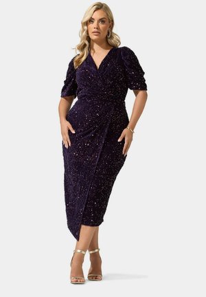 Femme blonde en robe portefeuille à sequins violet foncé avec manches bouffantes, debout, mains sur les hanches, portant des talons à brides dorées.