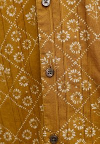 Ύφασμα μουστάρδας με λευκό γεωμετρικό μοτίβο λουλουδιών και κάθετη ραφή, με τρία καφέ κουμπιά με τέσσερις τρύπες σε κοντινή λήψη.