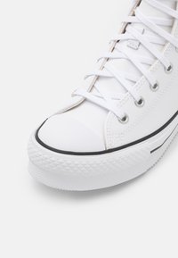 Gros plan d'une chaussure de toile blanche avec des lacets blancs, des œillets métalliques, un embout en caoutchouc et une semelle extérieure blanche texturée à bande noire.