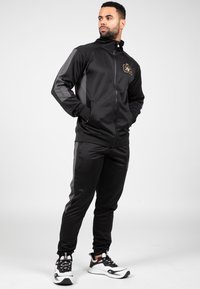 Zwarte sportieve trainingsoutfit van gladde stof, met een ritsjack en bijpassende broek met grijze zijaccenten. Inclusief logodetail.